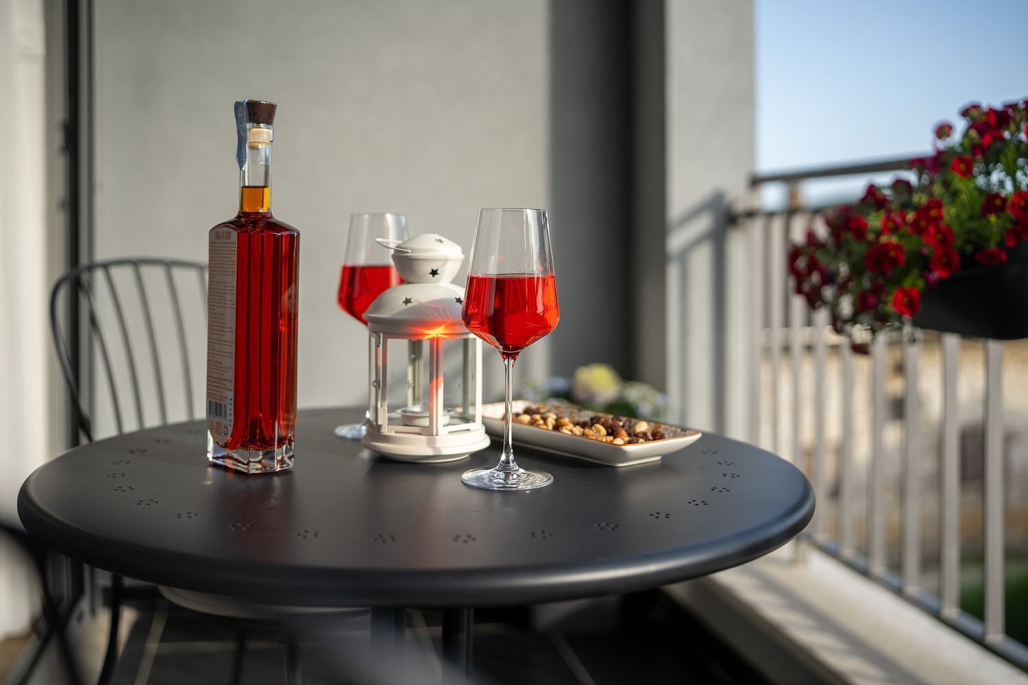 A table with a bottle, two glasses, a lantern, and a plate of nuts on a balcony.