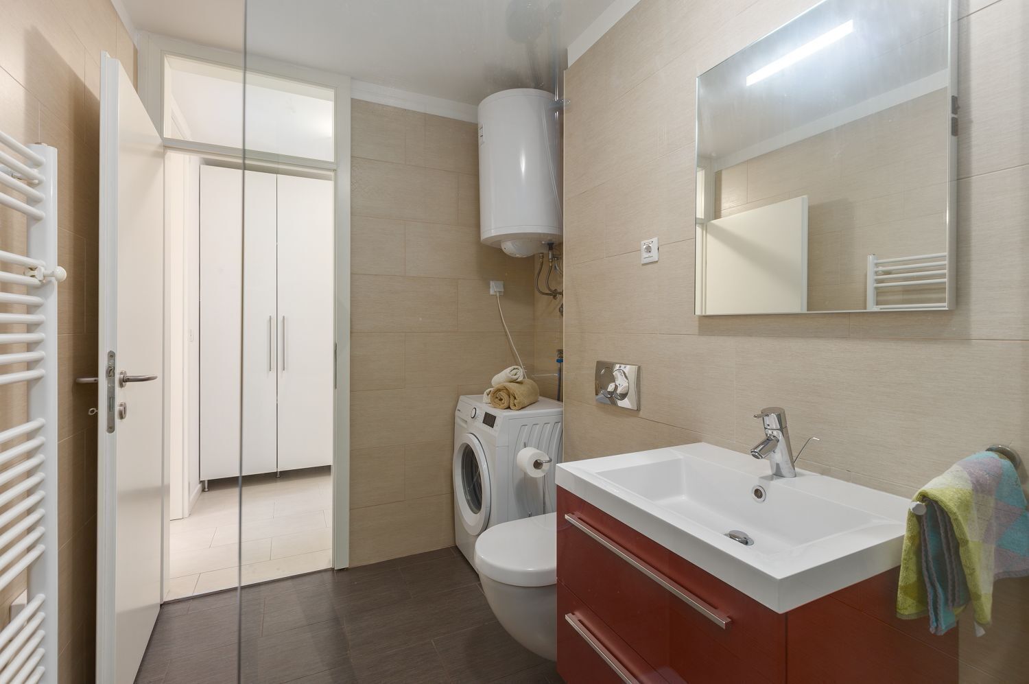 Une salle de bain moderne avec un lavabo blanc, un meuble rouge et un miroir au-dessus. On y trouve également un lave-linge et des toilettes dans un coin.