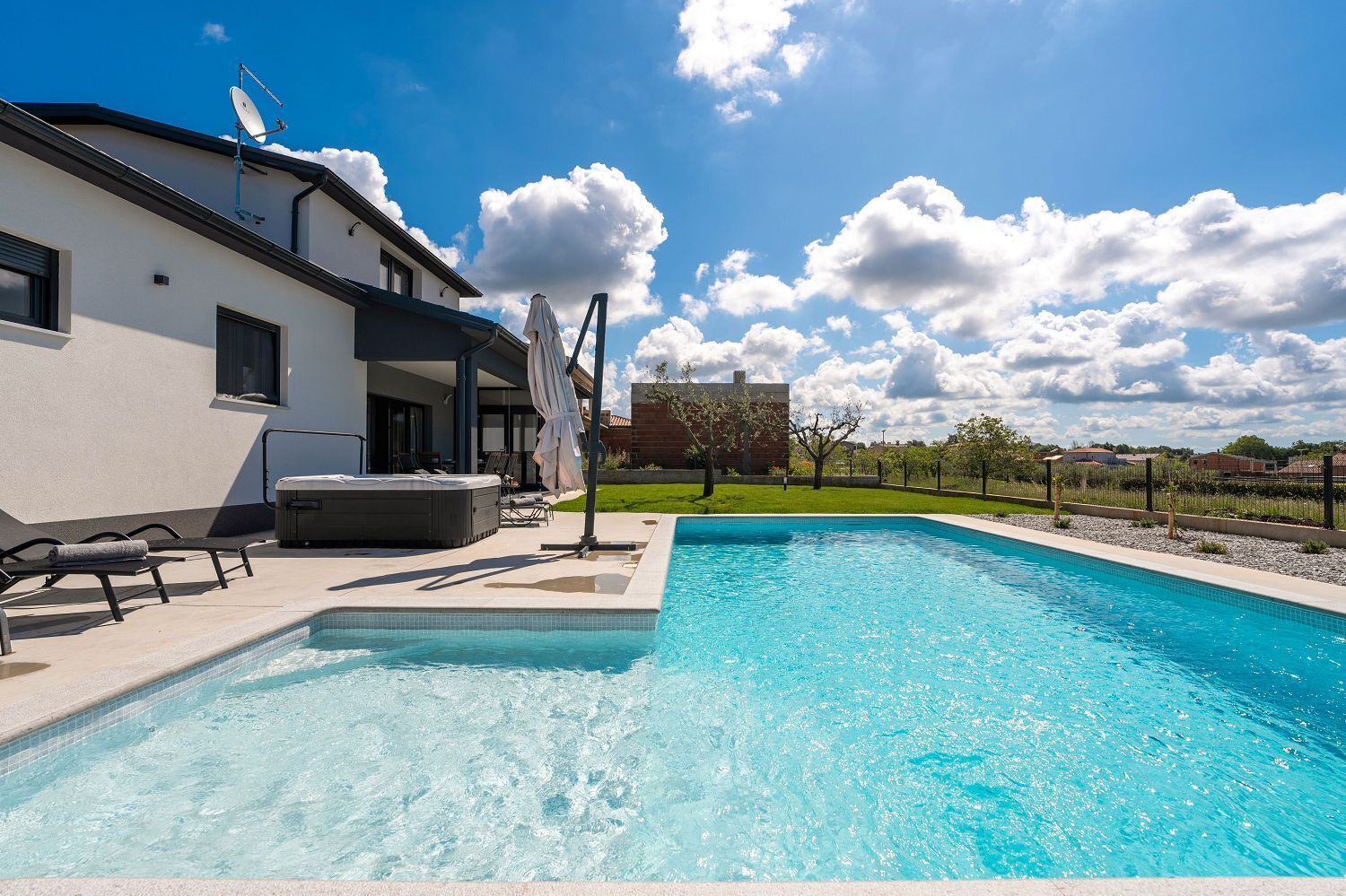 A swimming pool with lounge chairs and a hot tub, surrounded by a modern house and a clear sky.