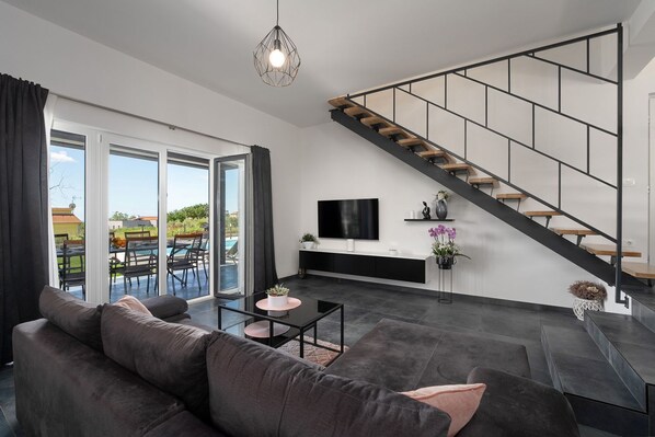 A modern living room with a dark gray sofa, a coffee table, and a TV mounted on the wall. A staircase with wooden steps leads to an upper level. - Impressive villa with sauna, private pool, whirlpool, panoramic sea view (KAŠTELIR)