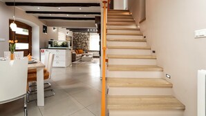 Un intérieur moderne avec un escalier en bois, un coin repas avec une table et des chaises blanches, et une cuisine dotée d’un mur en pierre et d’une cheminée en pierre.