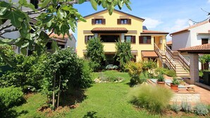 Una casa gialla a due piani con un balcone, immersa in un giardino curato con diverse piante e un piccolo prato.