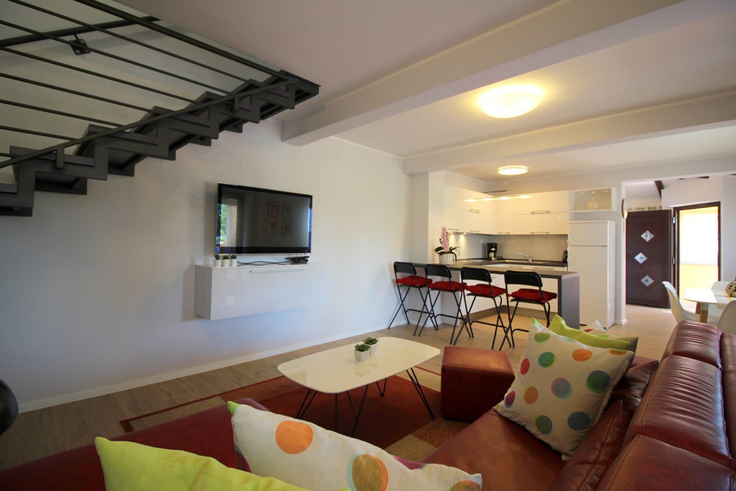 Ein modernes Wohnzimmer mit Flachbildfernseher, einer Essecke mit klappbaren Stühlen, einer Küche im Hintergrund und einer Treppe.