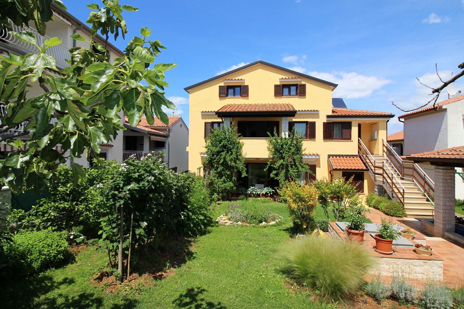 A two-story building with a yellow facade and a red-tiled roof, surrounded by a well-maintained garden with various plants and a grassy lawn.