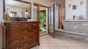 Une commode en bois avec des tiroirs, un miroir et un vase rempli de fleurs. On y trouve également une cheminée en pierre avec un rideau, ainsi qu’une fenêtre donnant sur un paysage verdoyant.