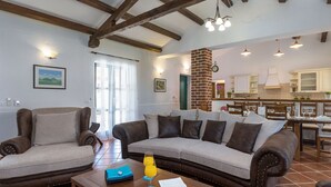 Una sala de estar con sofá, sillones y una mesa de centro. Se observa una cocina al fondo con mesas y sillas de comedor.