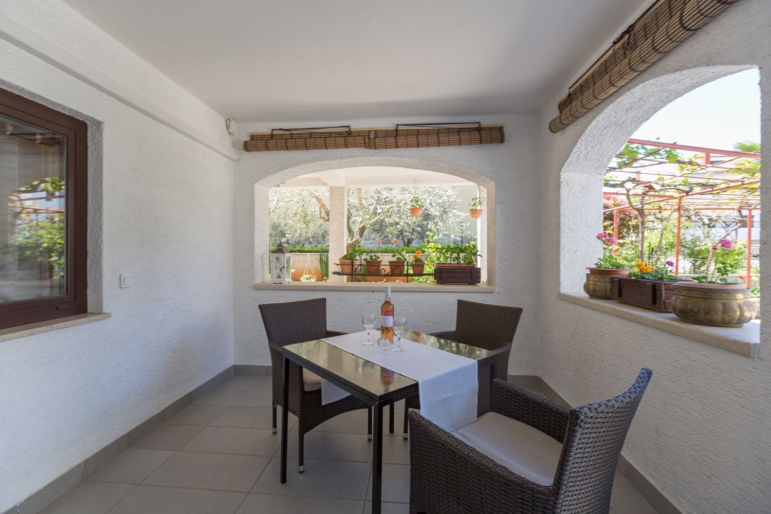 Ein kleiner, abgeschlossener Patio mit Tisch und Stühlen, ein Fenster mit Blumentöpfen und Blick auf einen Garten.