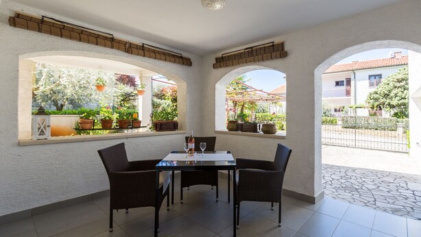 Un patio soleado con mesa y sillas, ventanas amplias con plantas en macetas, y vistas a una zona residencial.