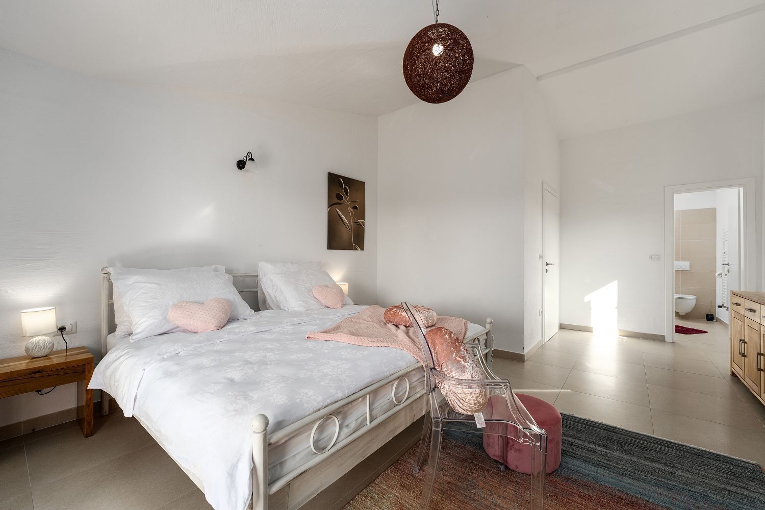 Une chambre à coucher comprenant un lit, des tables de chevet, un luminaire suspendu et une porte donnant sur une salle de bain.