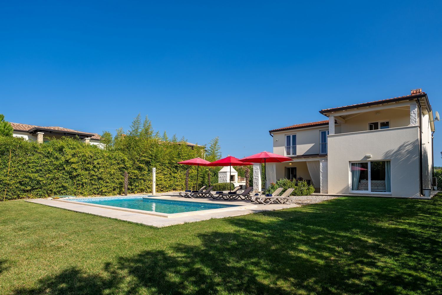Une maison à deux étages avec une piscine, des parasols rouges et une pelouse bien entretenue.
