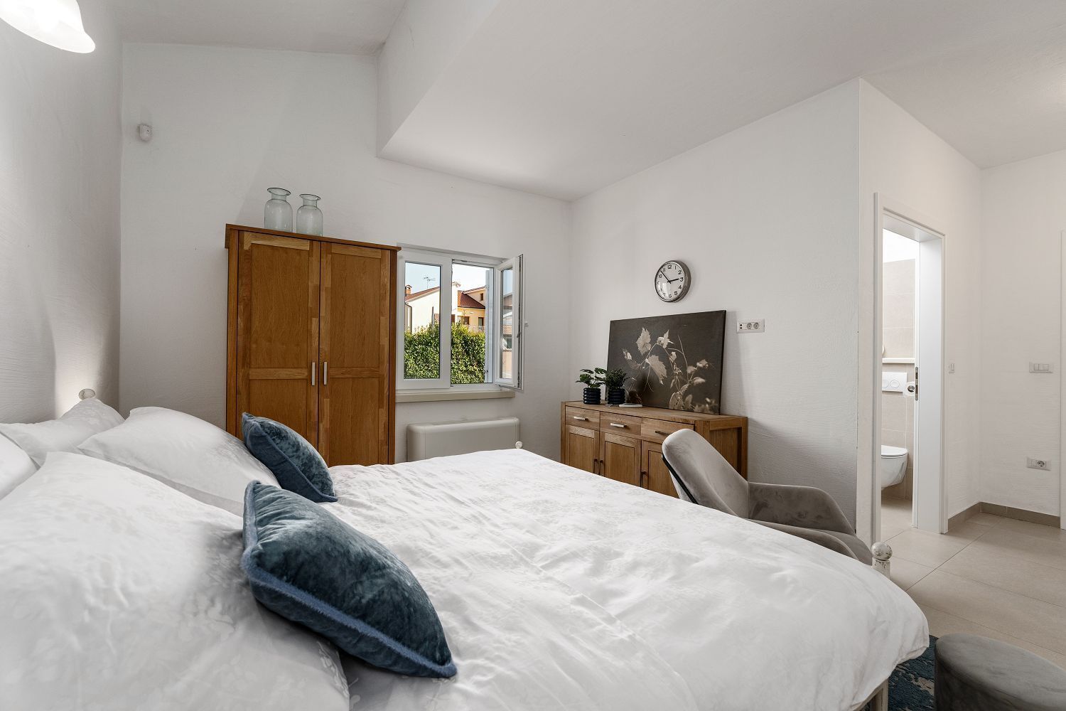 A bedroom with a large bed, wooden wardrobe, and a window with a view of buildings.