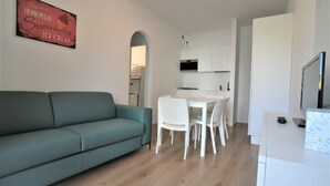 A modern living room with a teal sofa, a dining area with white chairs, and a kitchen with white cabinets. - Cozy Apartment With Balcony Near Piazza Torino (Jesolo)