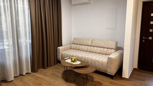 Una sala de estar moderna con un sofá color beige, una mesa de centro redonda de madera con un cuenco y flores, y un ventanal con cortinas traslúcidas.