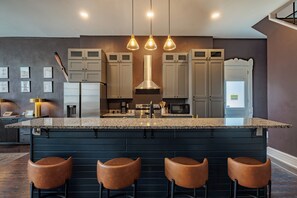 A modern kitchen with a central island, bar stools, and a dining area with a dark wood floor. - Arcade Game Room + Sleeps 12 Fast WiFi + Workspace Backyard Hangout (Dayton)
