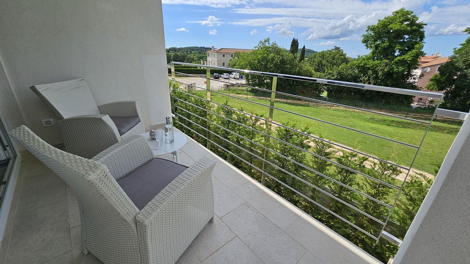 A balcony with wicker furniture, a glass table, and a view of a green landscape and buildings.