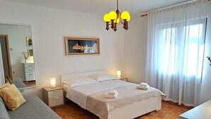 Un dormitorio ordenado con cama, mesitas de noche, un sofá, una lámpara de araña y una ventana con cortinas.