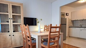 Un comedor con una mesa y sillas de madera, un televisor montado en la pared, y una zona de cocina con armarios y un refrigerador.