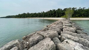 A rocky shoreline with a clear view of the water and a forested area in the background. - Private 3BR Gem on 2.5 Wooded Acres- Close to All (Sister Bay)
