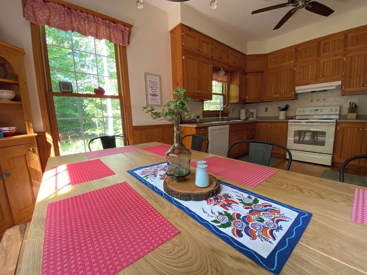 Une cuisine avec des meubles en bois, une table à manger avec des sets de table rouges, un vase rempli de fleurs et un ventilateur de plafond.