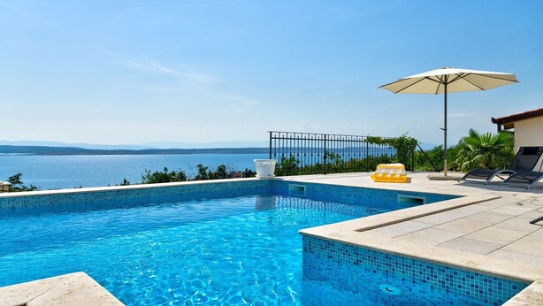 Una piscina con agua azul cristalina, vistas al mar y una zona de terraza con hidromasaje, sillones de descanso y una sombrilla.