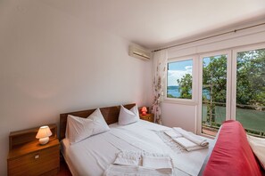 Ein Schlafzimmer mit einem großen Bett, einem Nachttisch mit Lampe, einem Fenster mit Blick auf Grün und Wasser sowie einer Klimaanlage an der Wand.