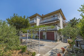 A two-story building with a balcony on the upper floor, a garage door, and a garden area with plants and flowers. - Charming apartment just 200 metres from the beach with parking, dishwasher and barbecue area (Jadranovo)