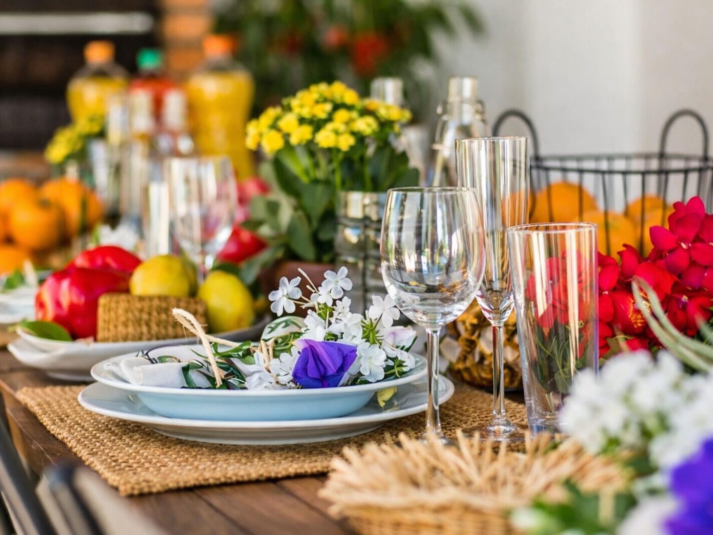 Ein Tisch ist gedeckt mit Tellern, Gläsern und Blumen, umgeben von Früchten und Flaschen.