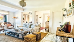 A spacious living room with a sofa, coffee table, and ottomans. There is a dining area with a table and chairs. The room has a large window and a chandelier. - Villa Alfredo in Estepona: 4 bedrooms near the beach (Estepona)
