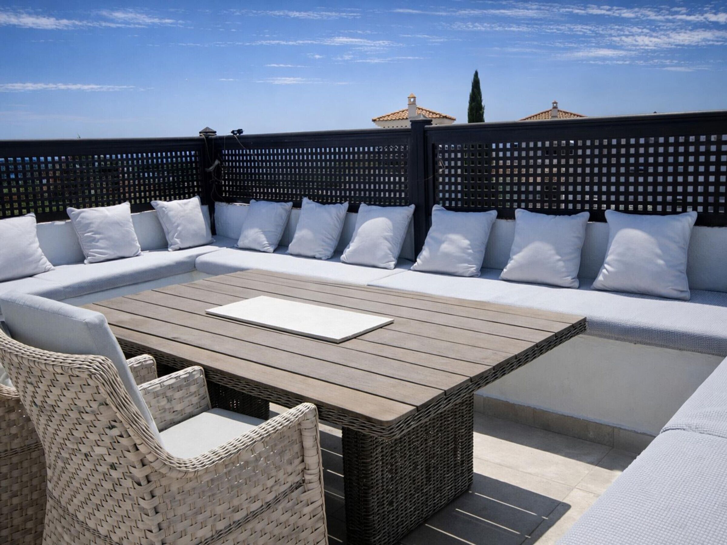Een terras op het dak met een houten tafel, rieten stoelen en een bank met witte kussens.