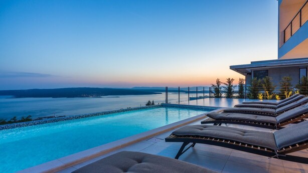 Ein moderner Infinity-Pool mit Liegestühlen und Blick auf das Meer im Dämmerlicht.