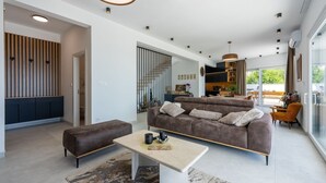 Ein modernes Wohnzimmer mit Sofa, Hocker und Couchtisch. Es gibt einen Essbereich mit Tisch und Stühlen. Der Raum hat ein großes Fenster und einen Blick ins Freie.