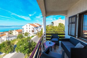 En balkon med udendørs møbler, et bord med en flaske og glas, samt udsigt til bygninger og havet.