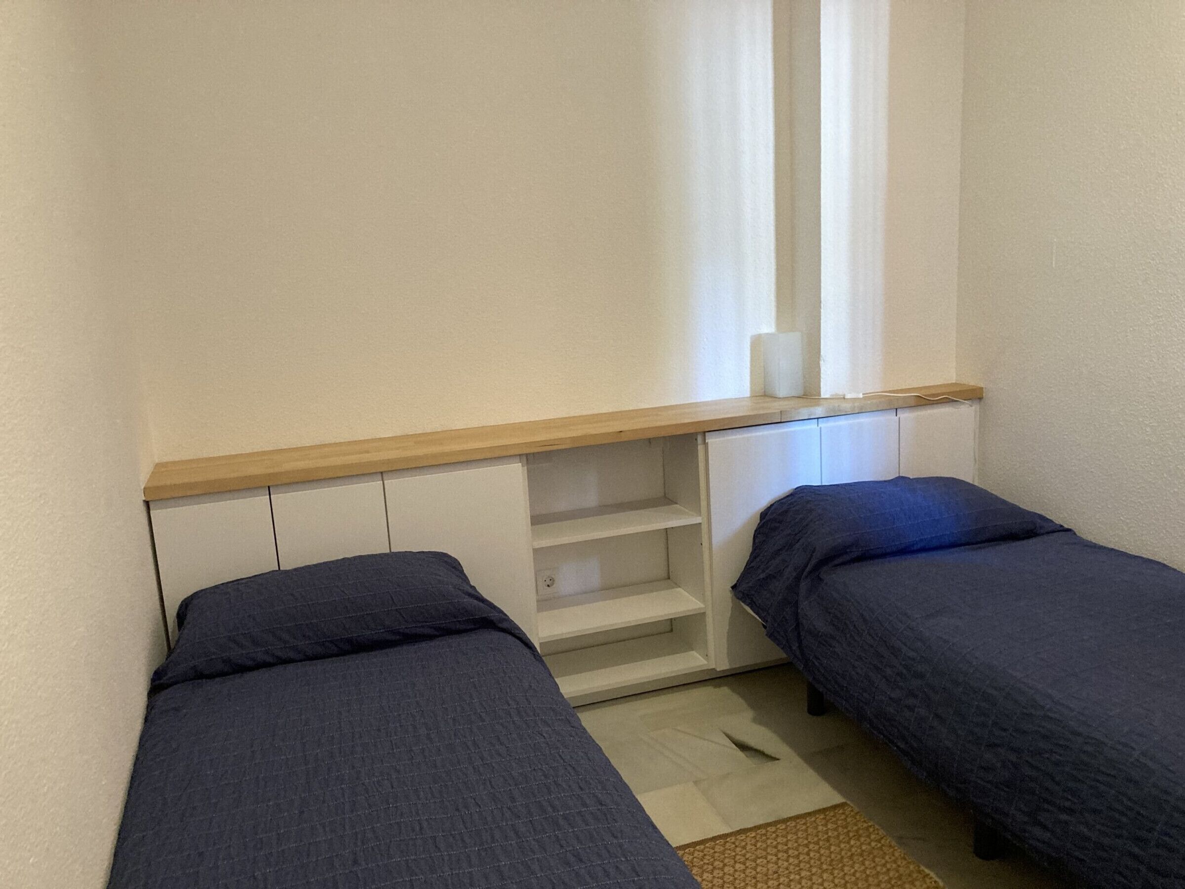 A small, compact room with two single beds, a wooden shelf, and a window with curtains.