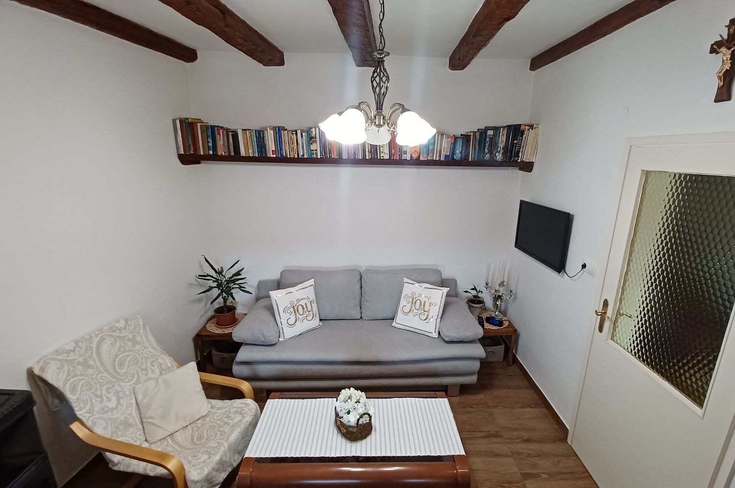 Un soggiorno con un divano grigio, una sedia di legno, un tavolino da caffè con una pianta, una libreria piena di libri, un lampadario e una porta con un pannello di vetro lavorato.