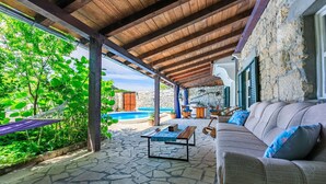Ein überdachter Außenbereich mit Steinboden, einem Pool, einem Holztisch und einer Couch mit Kissen.