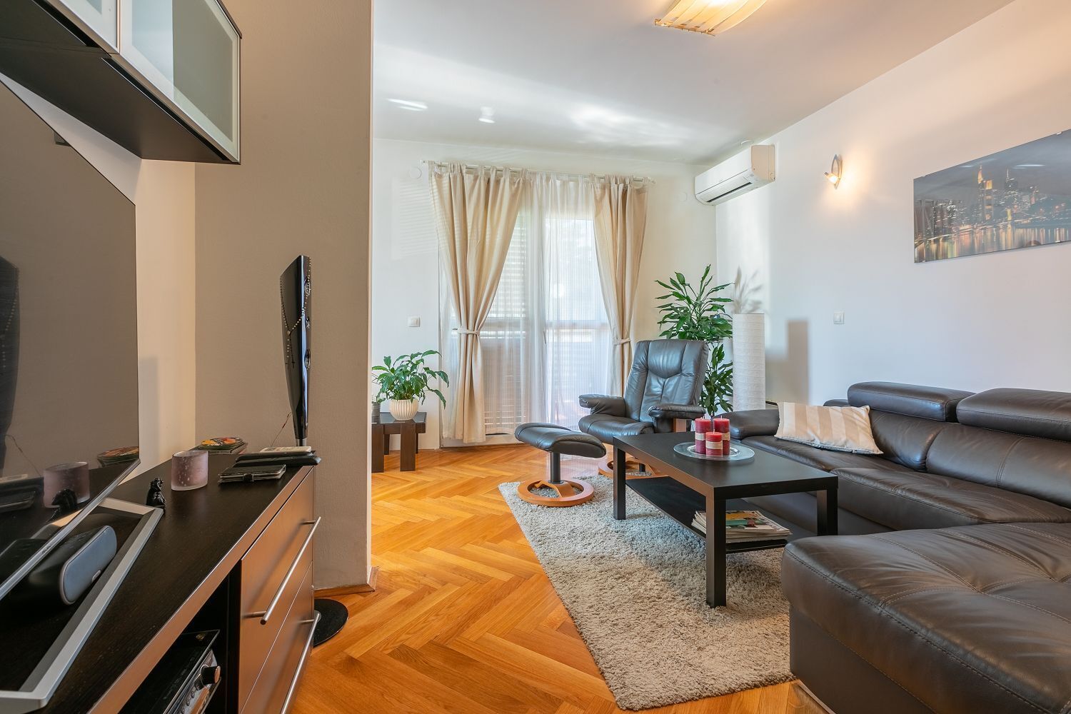 A modern living room with a sofa, coffee table, and TV. There is a window with curtains and a plant on the table.