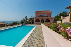 Ein Swimmingpool mit Fliesen umgeben, ein rosafarbenes Haus mit Rundbogenfenstern und Blick auf das Meer.