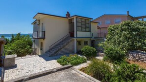 Edificio de dos pisos con una escalera de piedra que lleva a la entrada, rodeado de vegetación y una vista despejada al mar.
