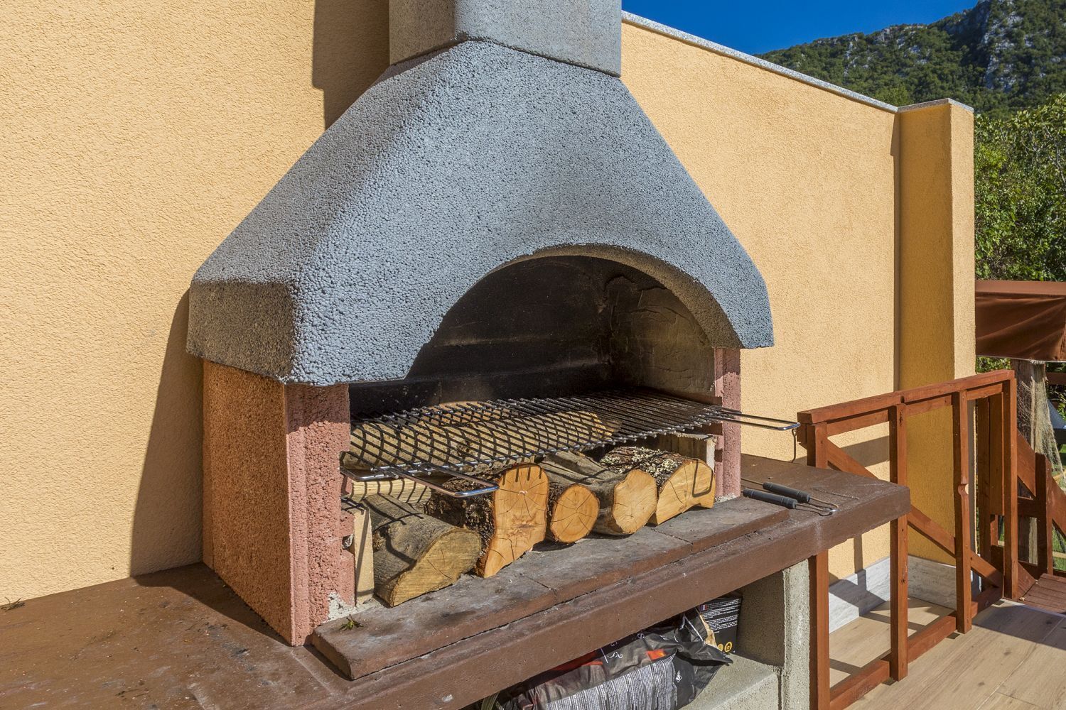 An outdoor stone-built pizza oven with a chimney, wood logs, and a metal grill rack.