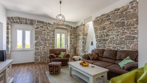 A cozy living room with stone walls, a brown leather sofa, a matching armchair, a wooden coffee table with fruit, and a colorful ottoman. - Unique villa with private pool, sea view, trampoline, bikes and many hiking trails in the area (Jadranovo)