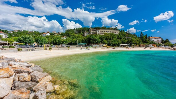 A coastal beach with clear turquoise waters, a rocky shoreline, and buildings in the background. - Unique villa with private pool, sea view, trampoline, bikes and many hiking trails in the area (Jadranovo)
