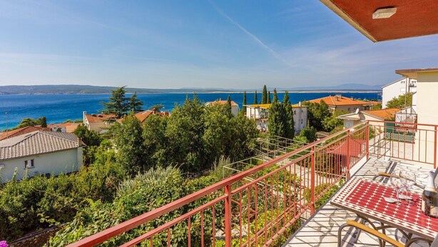 Ein Balkon mit roter Brüstung bietet Blick auf die Küste mit Häusern und Bäumen.