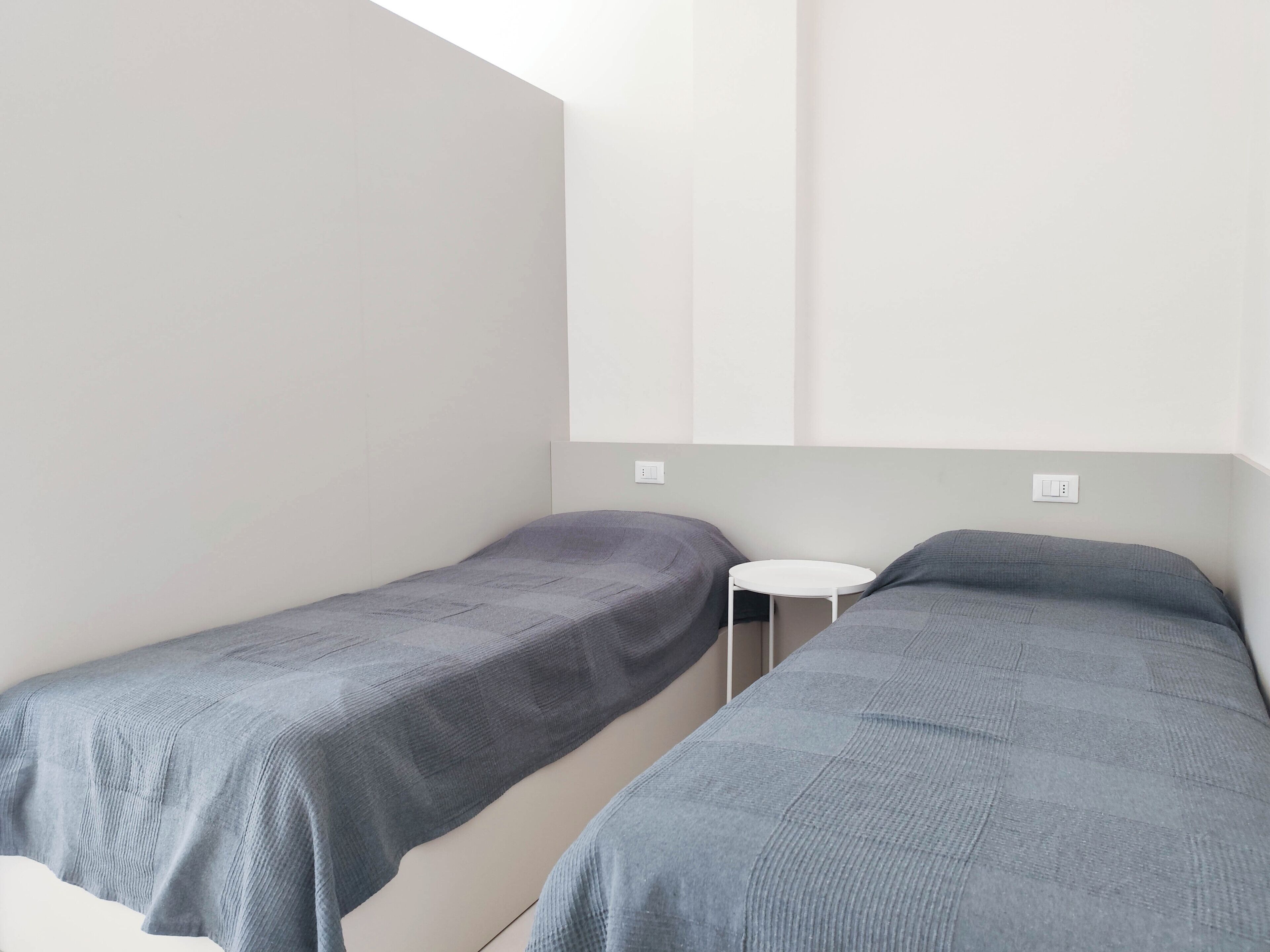 A small, minimalist room with two single beds, a small white table, and a simple, clean design.