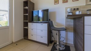 Ein gemütlicher Wohnraum mit Fernseher auf einem Holzschrank, einem kleinen Barhocker und einer Küchenzeile mit Kaffeemaschine.