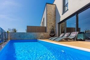 Piscina moderna al aire libre con pared de piedra y cubierta de madera, rodeada por una cerca metálica y una silla colgante.