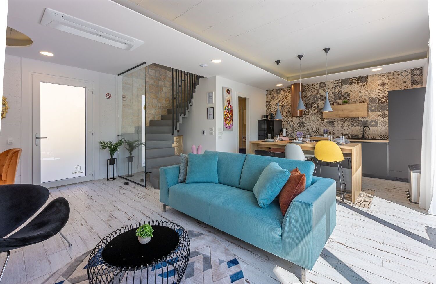 Un salon moderne avec un canapé bleu, une table ronde noire agrémentée d’une plante, un escalier, un coin cuisine et une table à manger avec des chaises.