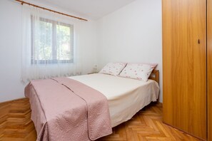 Ein Schlafzimmer mit einem Bett, einem Fenster mit durchscheinenden Vorhängen, einem Holzschrank und einem Parkettboden mit Fischgrätenmuster.