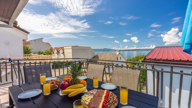 A table set with breakfast items including fruits, cheese, and meats, overlooking a scenic view of the sea and mountains.