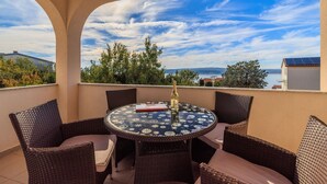 Un tavolo rotondo con sedie in vimini su un balcone con vista libera sul mare e sugli alberi.