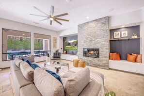A modern living room with a stone fireplace, a ceiling fan, and a seating area with cushions. - Carefree Casa: Resort-Style Home w/Heated Pool+Spa, Arcade, Pizza Oven & Casita (Indio)